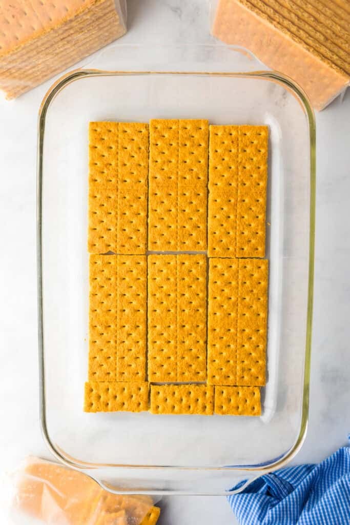A layer of graham crackers arranged neatly in a glass baking dish, ready to become the base for a delicious lemon eclair icebox cake.