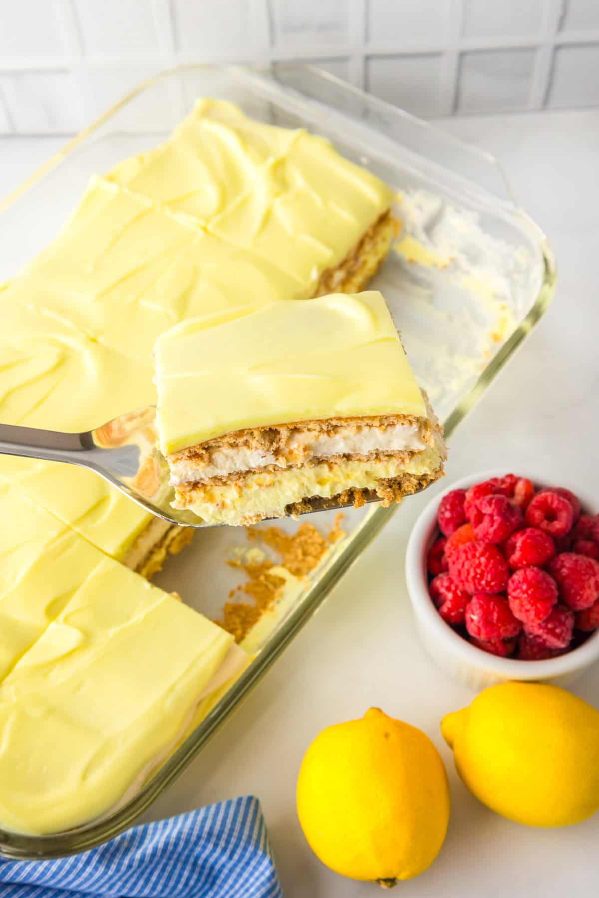 A glass baking dish with a sliced no bake lemon eclair cake topped with creamy frosting, a metal spatula holding one piece.