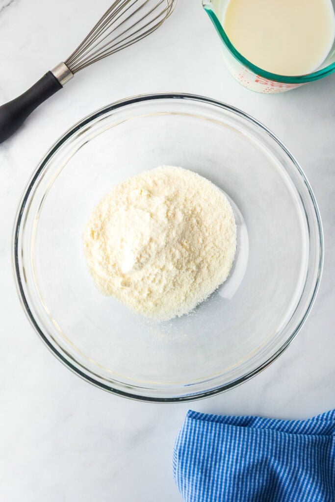A glass mixing bowl with instant lemon pudding mix, a whisk, a measuring cup of milk, and a blue striped cloth on a counter for making a lemon eclair icebox cake.