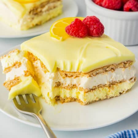 A slice of no bake lemon eclair cake with creamy layers and graham crackers, topped with a lemon slice and raspberries.