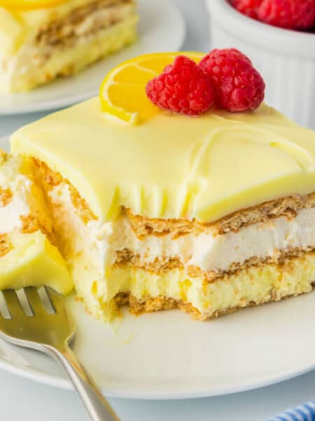 A slice of no bake lemon eclair cake with creamy layers and graham crackers, topped with a lemon slice and raspberries.