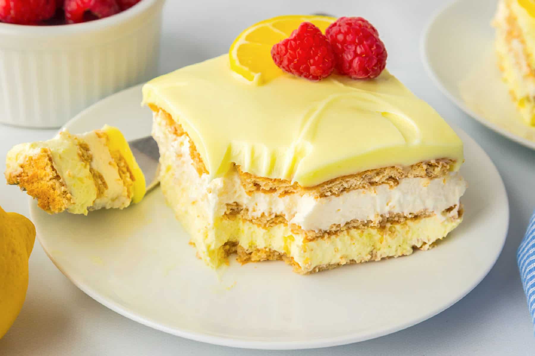 A slice of layered lemon eclair icebox cake topped with lemon glaze and berries sits on a plate, with a forkful removed.