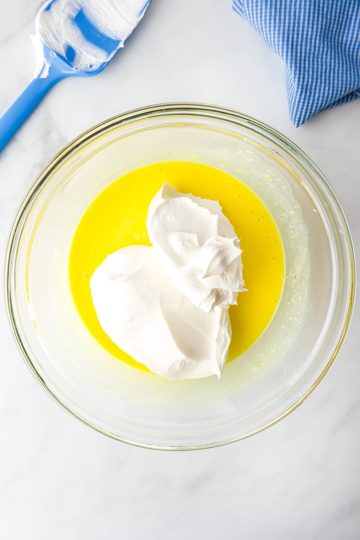 A glass bowl with yellow lemon pudding and dollops of Cool Whip being mixed together for a layer of lemon eclair icebox cake.
