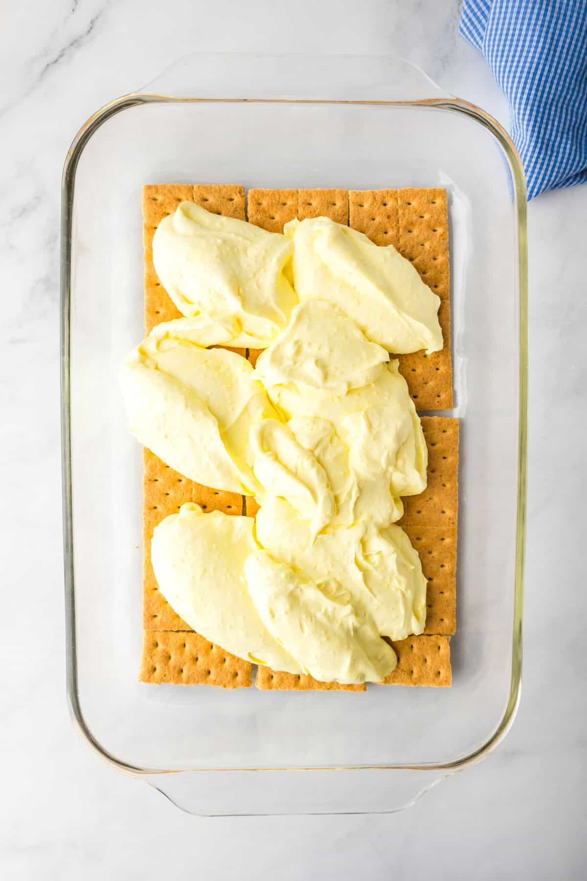 A glass baking dish with a layer of graham crackers and dollops of cheesecake puffing being smoothed for a lemon eclair icebox cake.