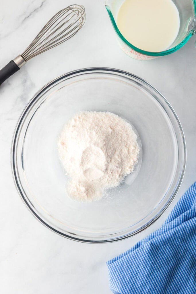 A glass mixing bowl full of dry instant cheesecake pudding mix for a lemon eclair icebox cake, a whisk and a measuring cup of milk nearby.