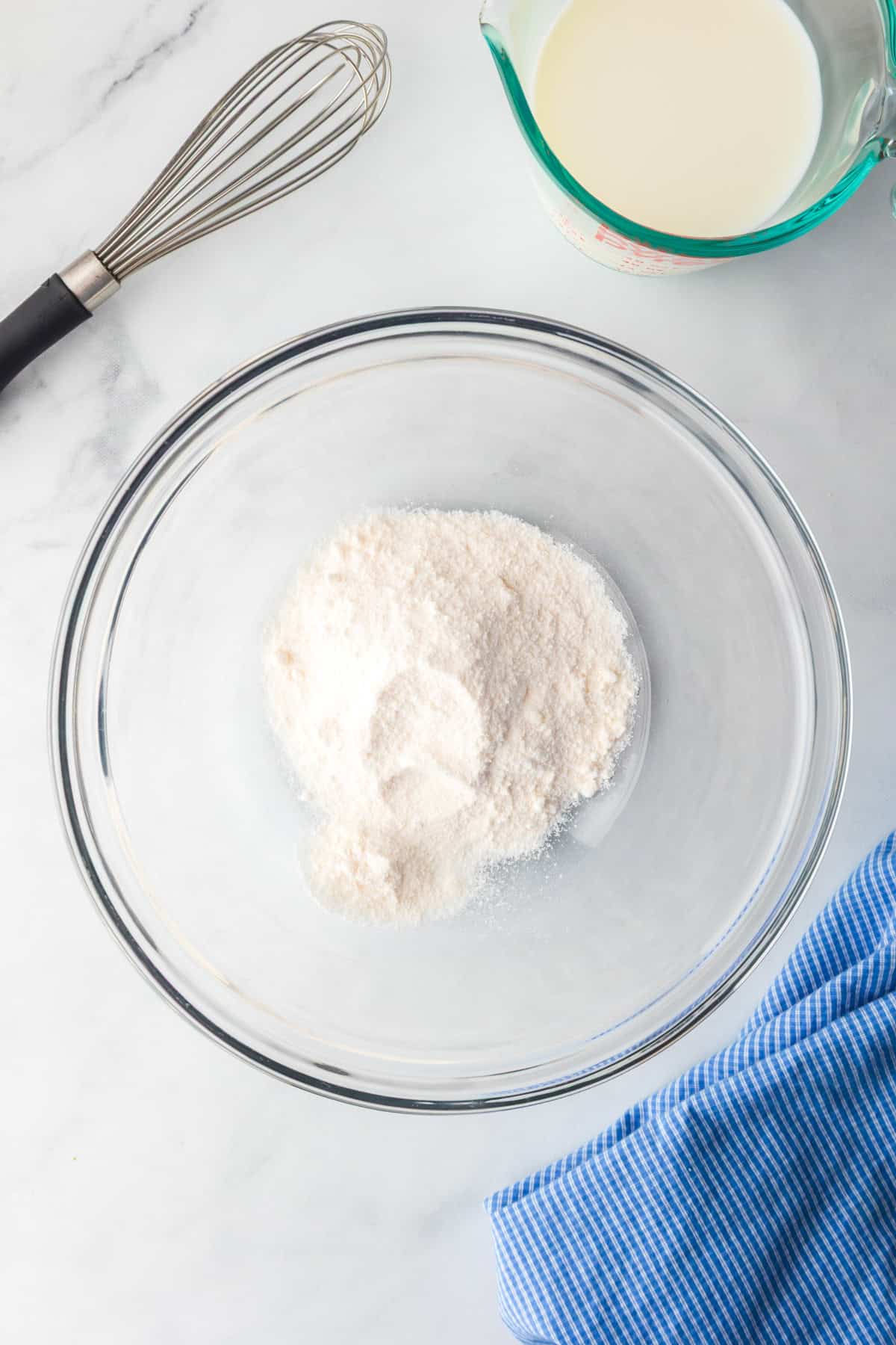 A glass mixing bowl full of dry instant cheesecake pudding mix for a lemon eclair icebox cake, a whisk and a measuring cup of milk nearby.