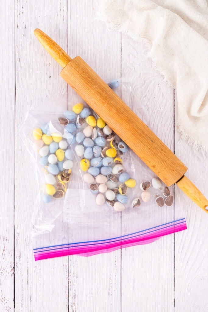 A rolling pin rests on a sealed plastic bag filled with colorful candy-coated chocolates, ready to be crushed for delicious mini egg chocolate chip cookies on a white wooden surface.