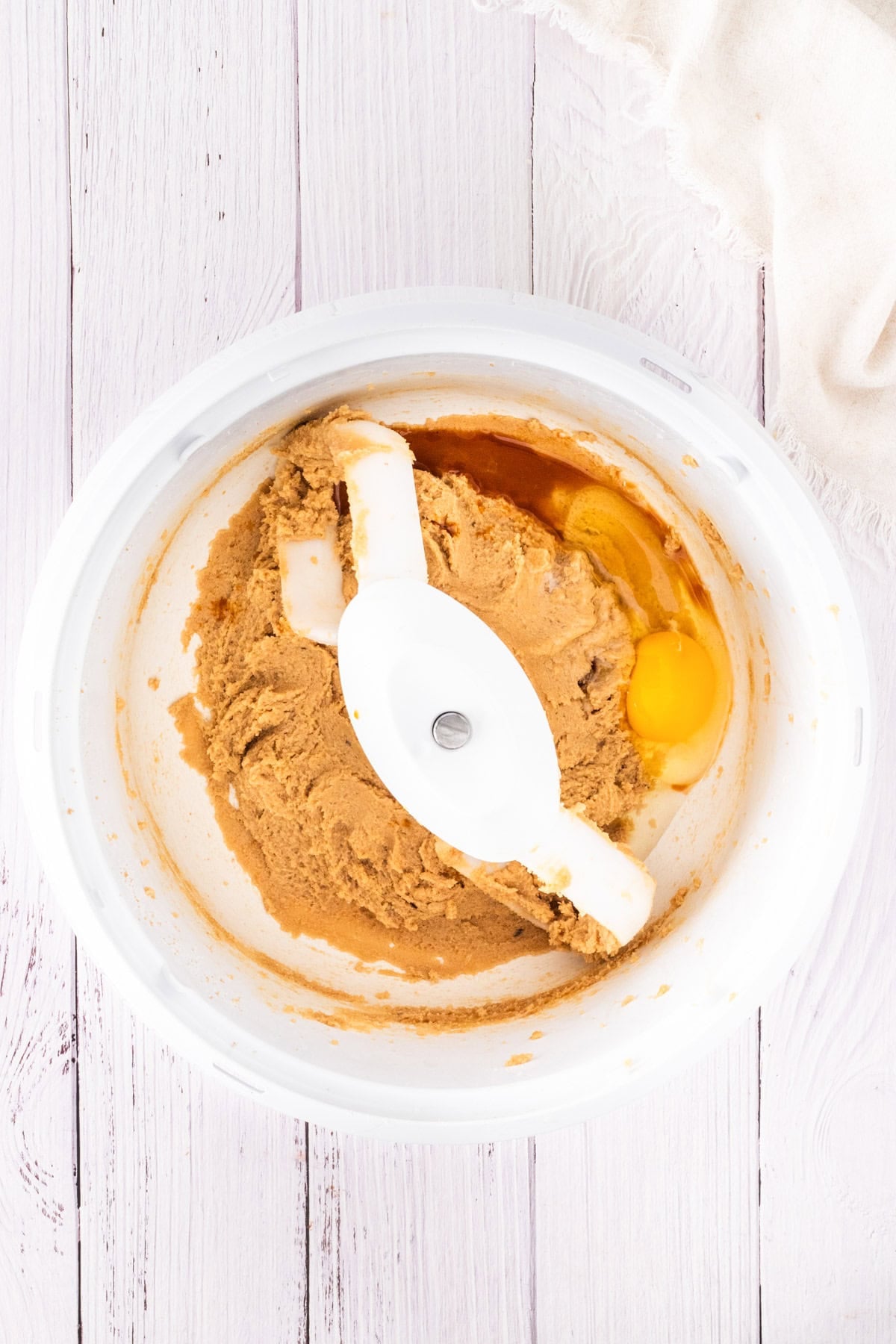 A mixing bowl with brown sugar, butter, an egg, and vanilla being combined for mini egg chocolate chip cookies with a white paddle attachment on a white wooden surface.