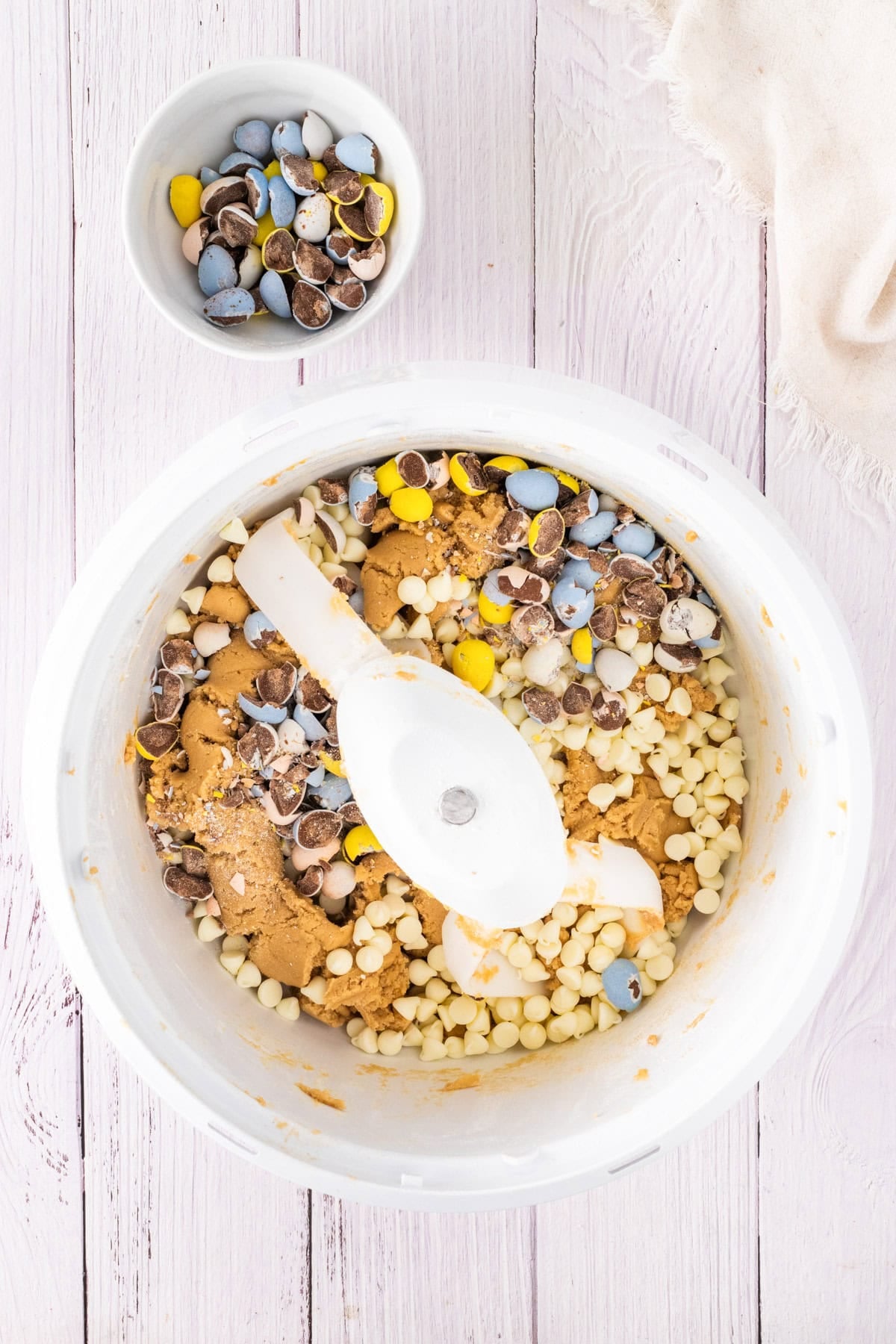 A mixing bowl filled with cookie dough, white chocolate chips, and crushed mini egg chocolate chip cookies sits next to a small bowl of more crushed candy eggs on a white wooden surface.