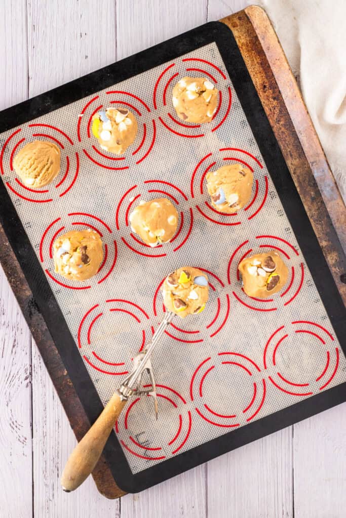 A baking sheet with round mini egg chocolate chip cookies on a silicone mat, some topped with small chocolate eggs. A cookie scoop with dough rests on the tray.