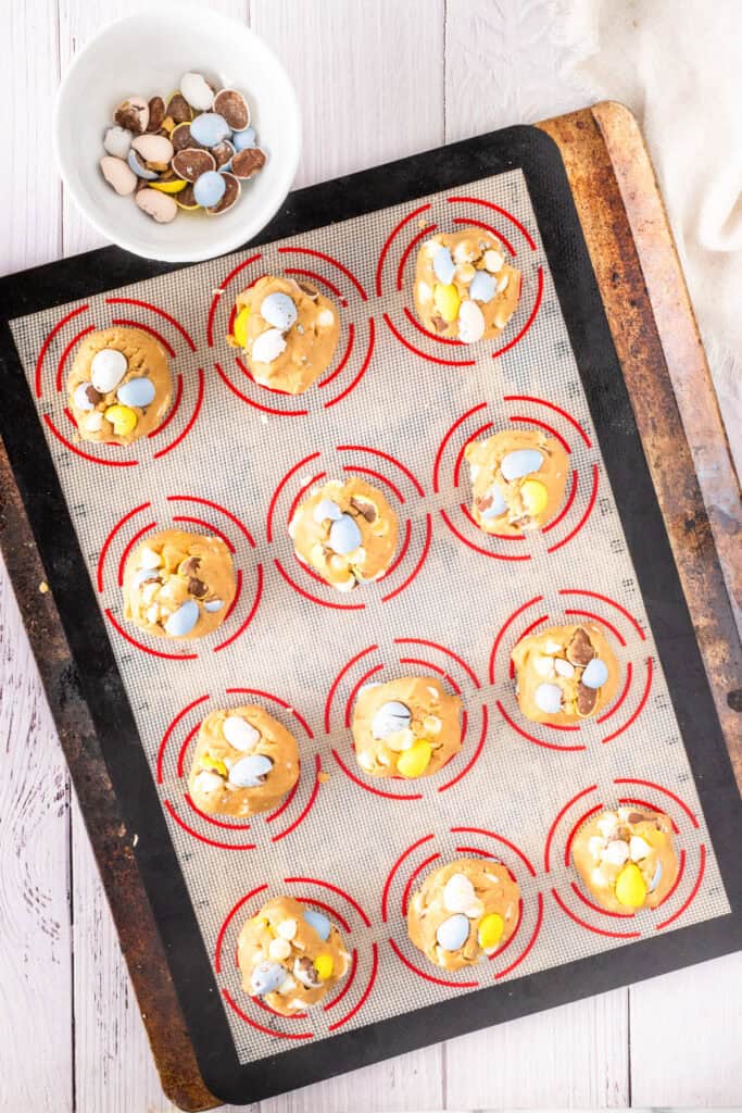 A baking sheet with twelve unbaked mini egg chocolate chip cookies, each topped with colorful chocolate eggs, sits on a white and red silicone mat. A small bowl of extra chocolate eggs is nearby.