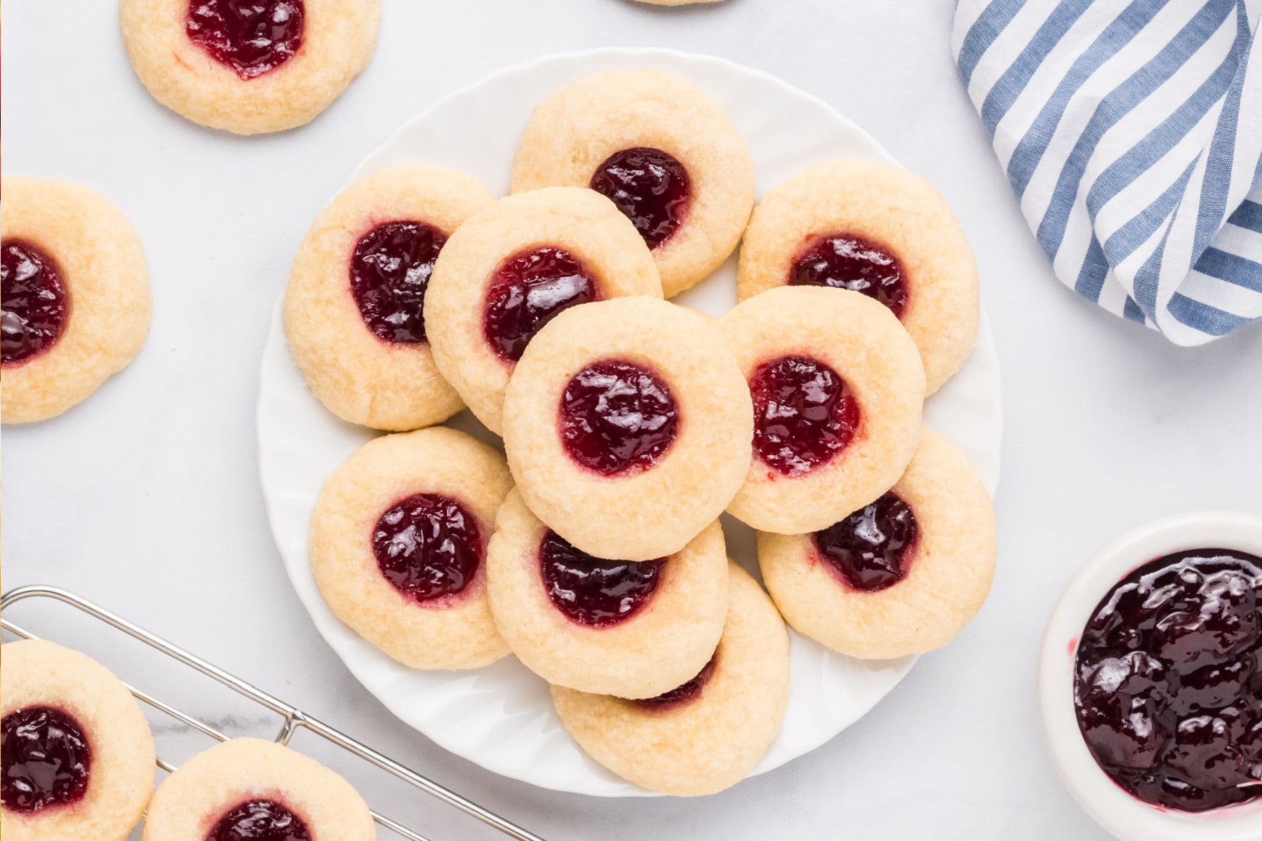 A plate of raspberry thumbprint cookies with jam centers, surrounded by more cookies, a bowl of jam, and a blue and white striped cloth.