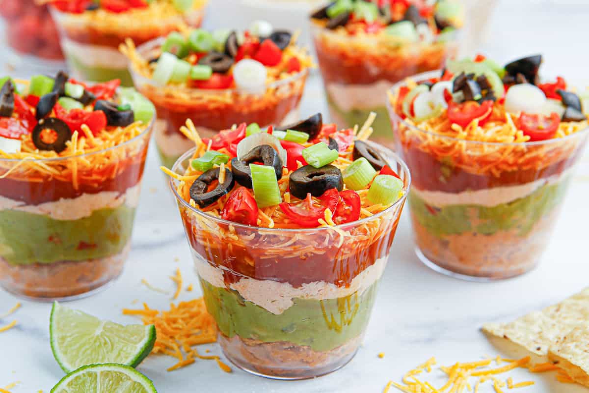 Individual clear cups filled with seven layer dip, including beans, guacamole, sour cream, salsa, shredded cheese, diced tomatoes, olives, and green onions.