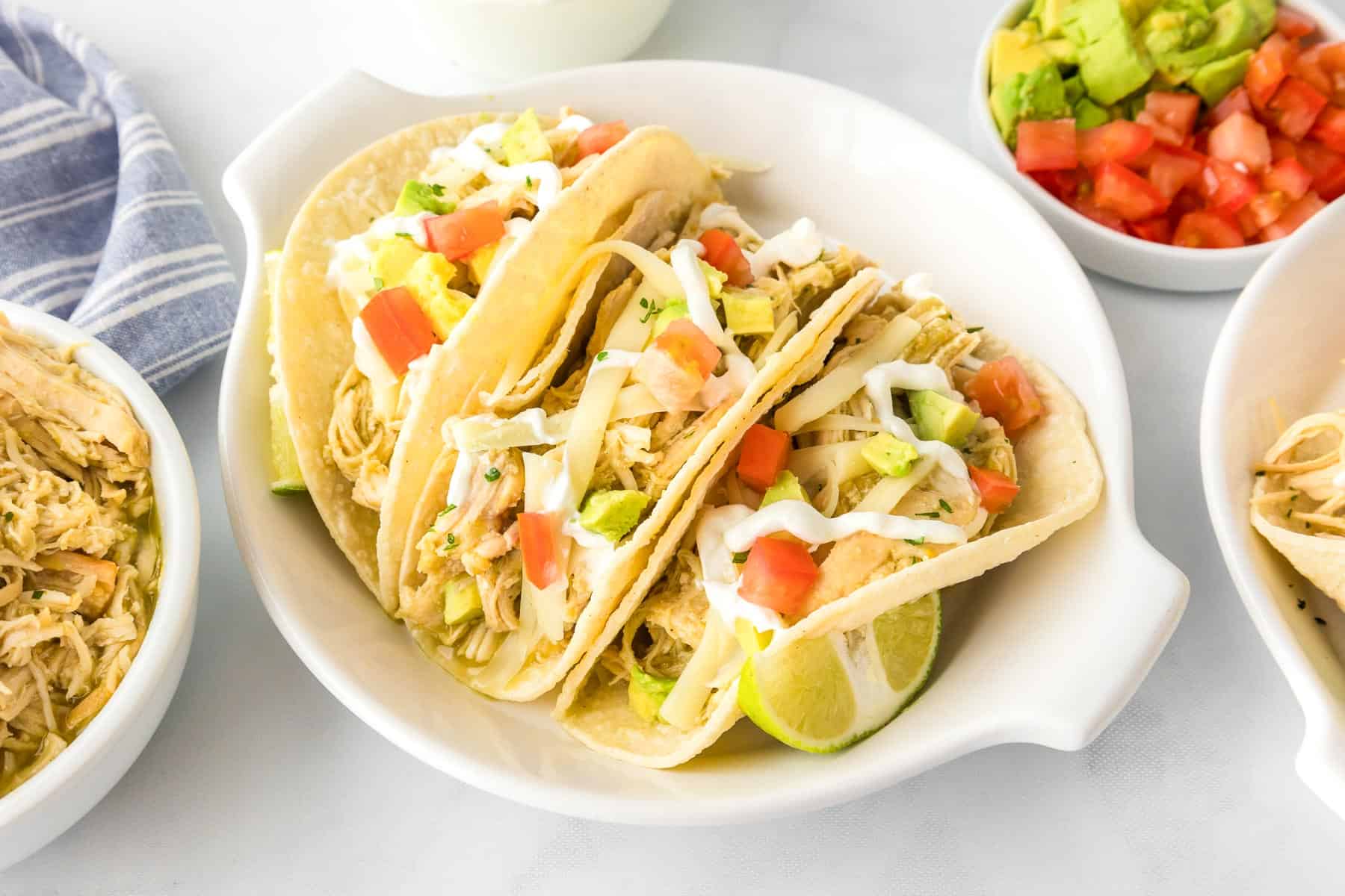 A white dish holds three Slow Cooker Salsa Verde Chicken tacos topped with shredded cheese, diced tomatoes, avocado, and sour cream, with lime wedges on the side.