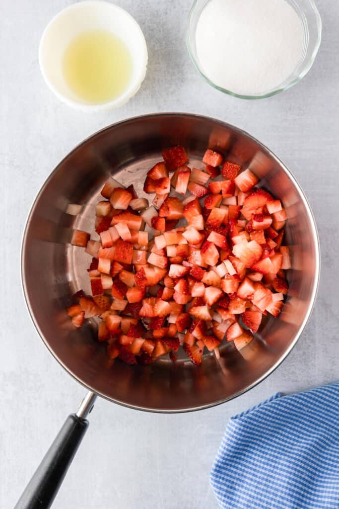A saucepan with chopped strawberries sits on a countertop next to bowls of lemon juice and sugar, ready for making a delicious strawberry jello cake.