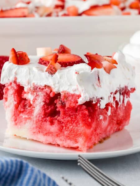 A slice of strawberry jello cake topped with whipped cream and fresh strawberry pieces sits on a white plate with a fork, with a casserole dish full of more strawberry jello cake in the background.