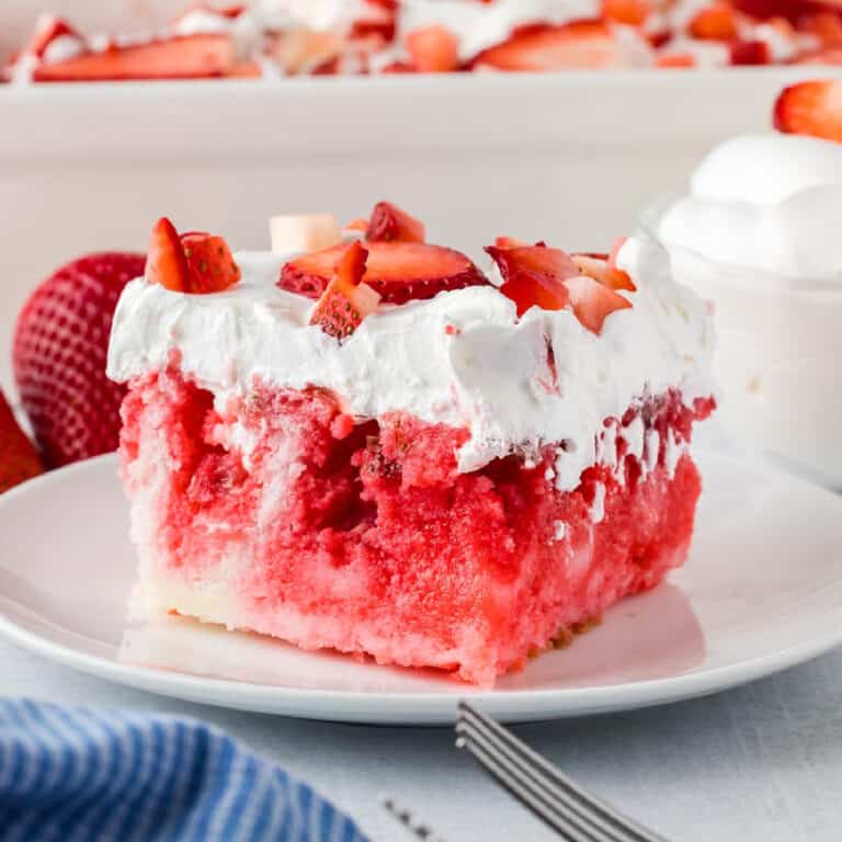 A slice of strawberry jello cake topped with whipped cream and fresh strawberry pieces sits on a white plate with a fork, with a casserole dish full of more strawberry jello cake in the background.