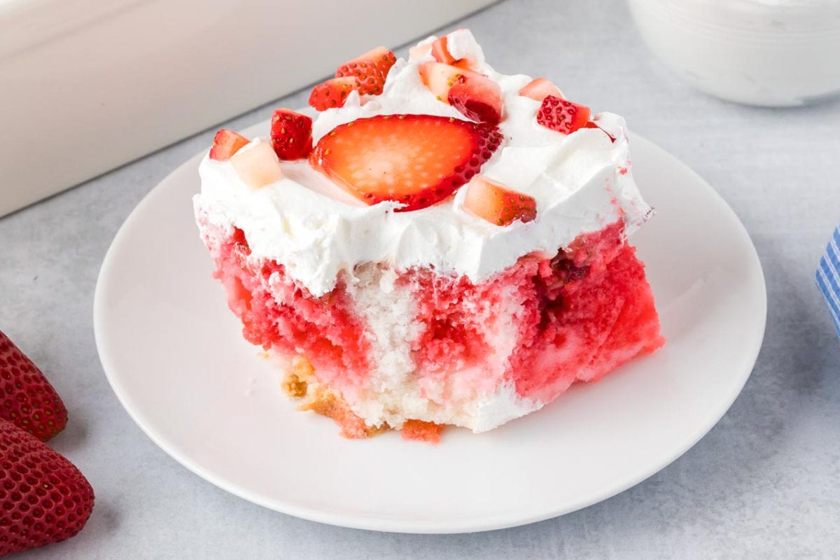 A slice of strawberry jello cake with whipped topping and fresh strawberry pieces on a white plate with a bite missing.