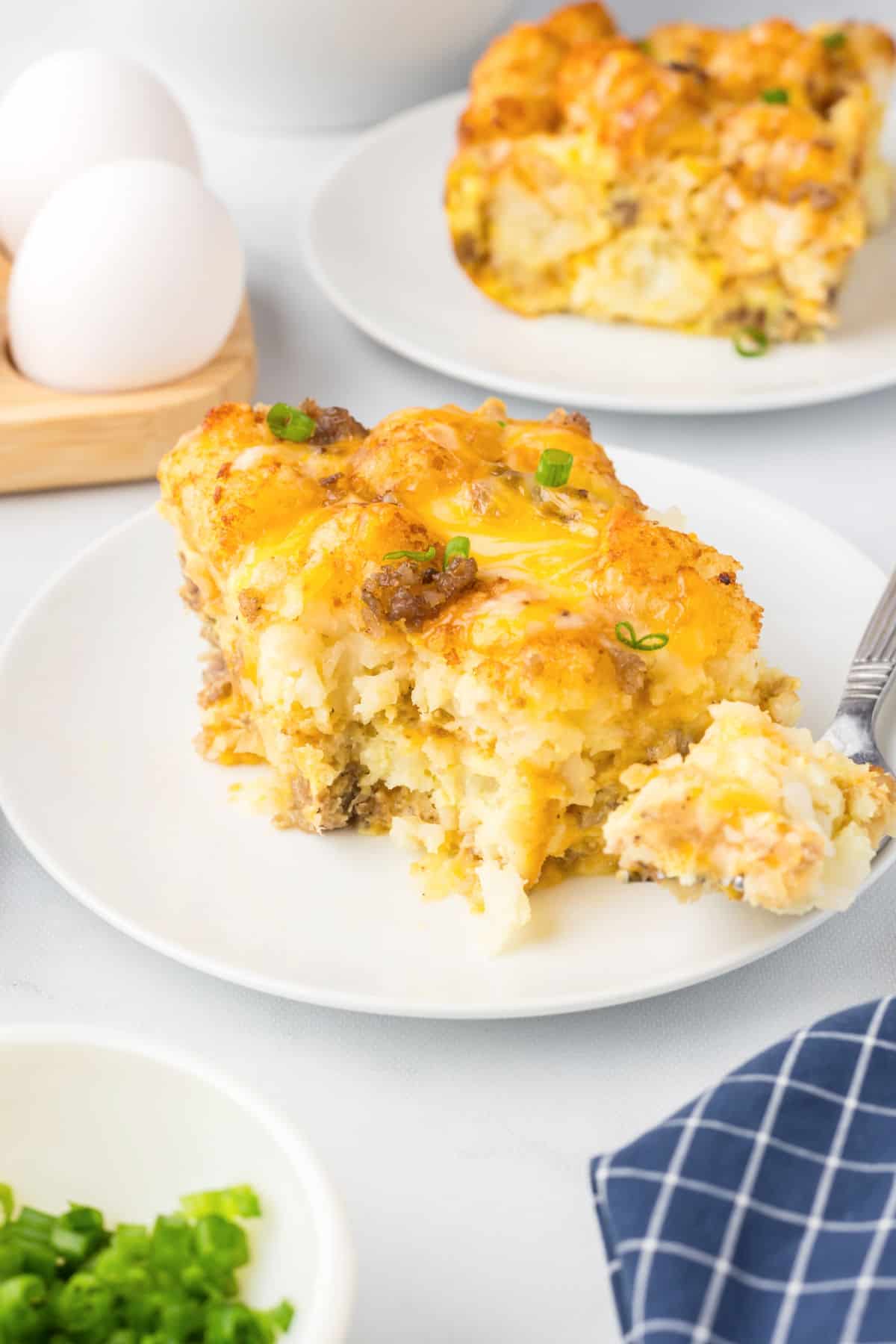 A slice of cheesy tater tot sausage breakfast casserole with ground meat and potatoes is served on a white plate; a forkful is taken out, and eggs and chopped green onions are nearby.