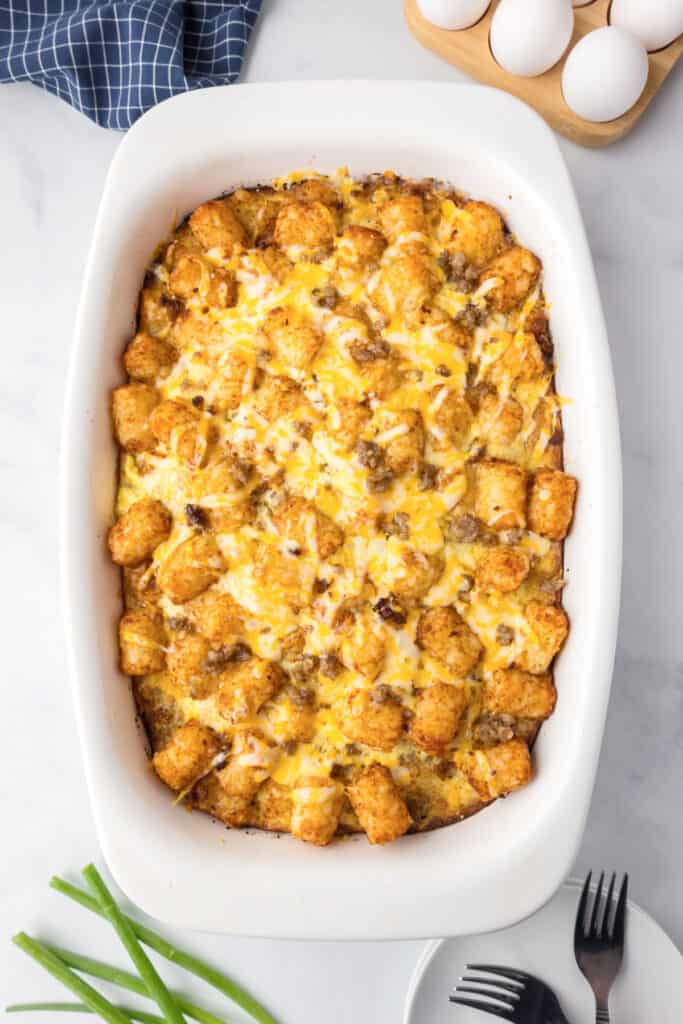 A tater tot breakfast casserole in a white dish, topped with melted cheese and sausage crumbles. Eggs, a napkin, green onions, and utensils are nearby.