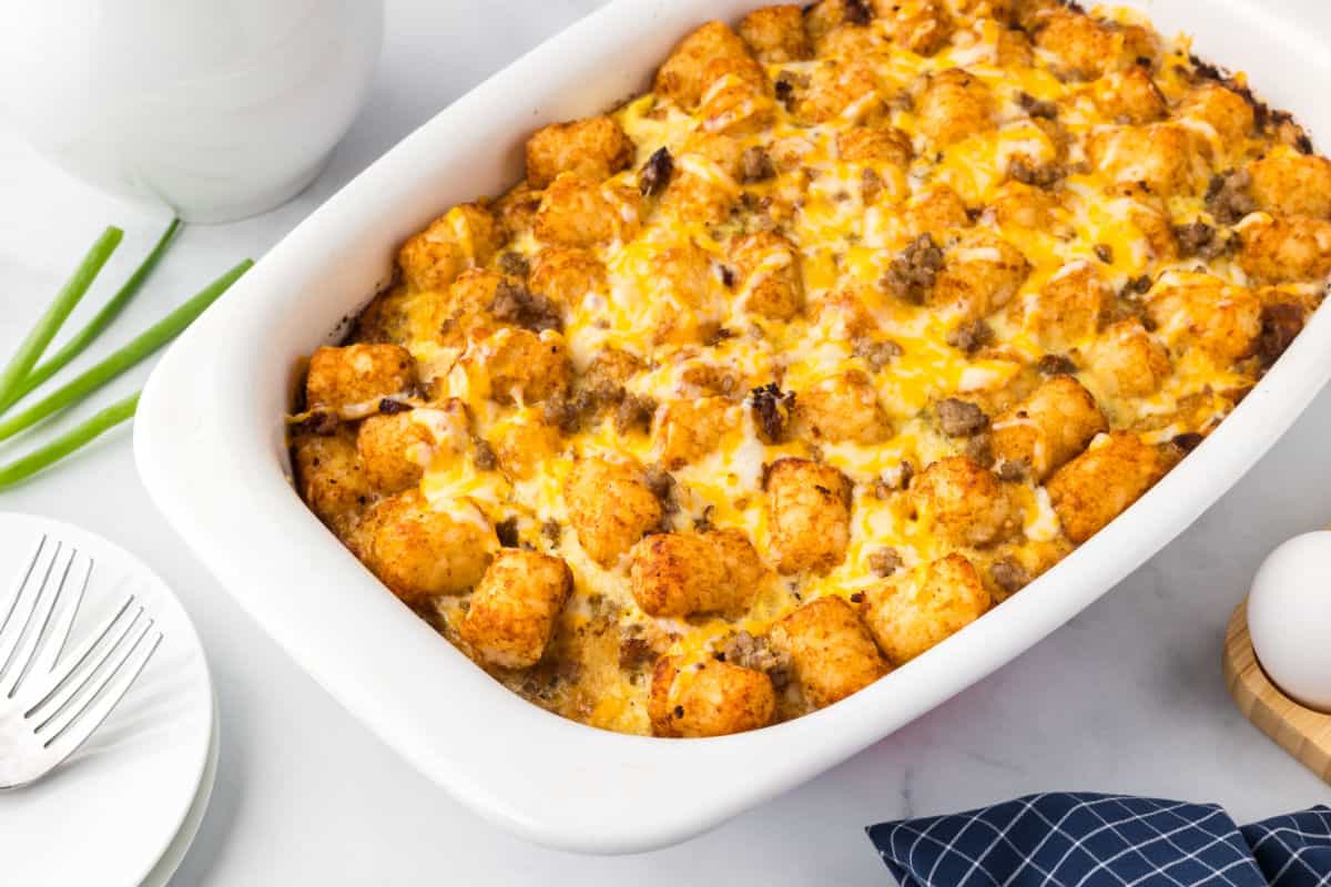 A baked casserole topped with golden tater tots, melted cheese, and pieces of sausage in a white baking dish, with green onions, forks, and an egg nearby on the table.