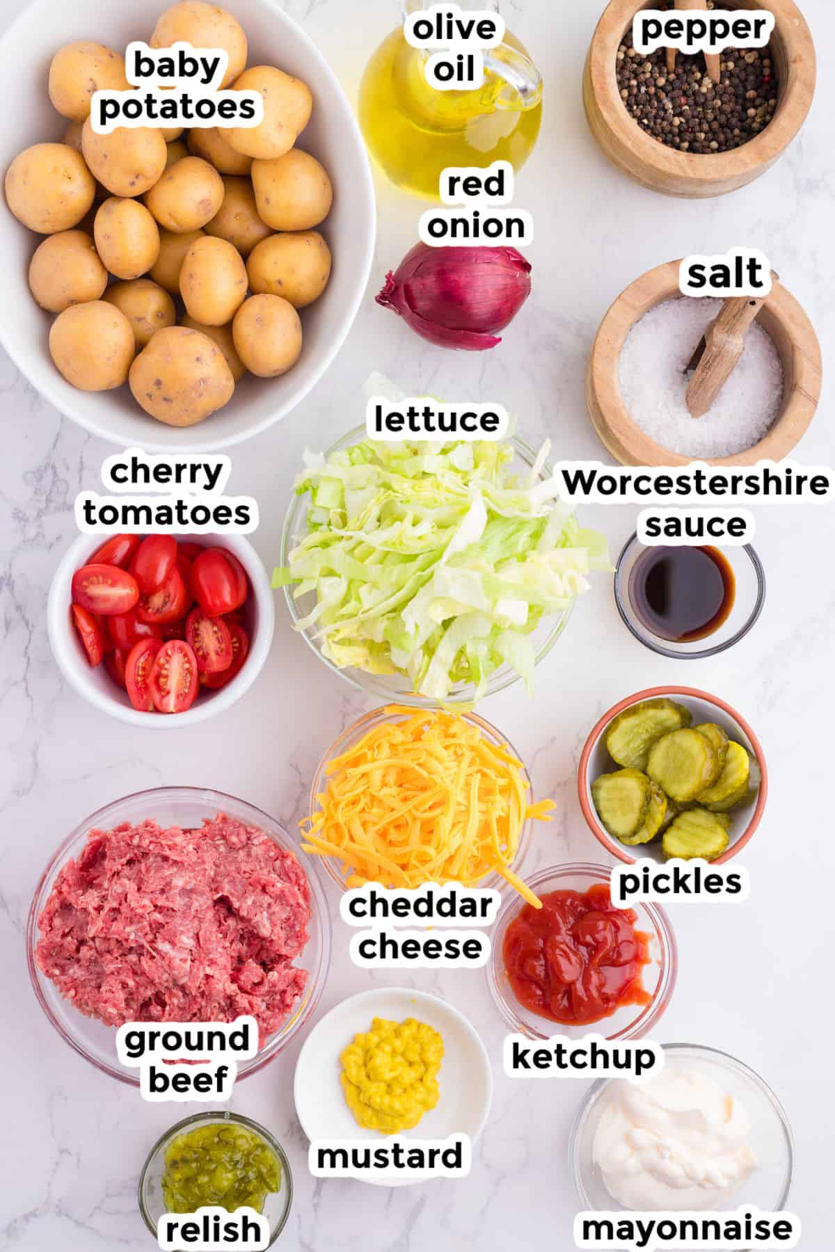 Assorted burger bowl ingredients on a white surface, including baby potatoes, ground beef, lettuce, cherry tomatoes, pickles, cheddar cheese, red onion, and various condiments.
