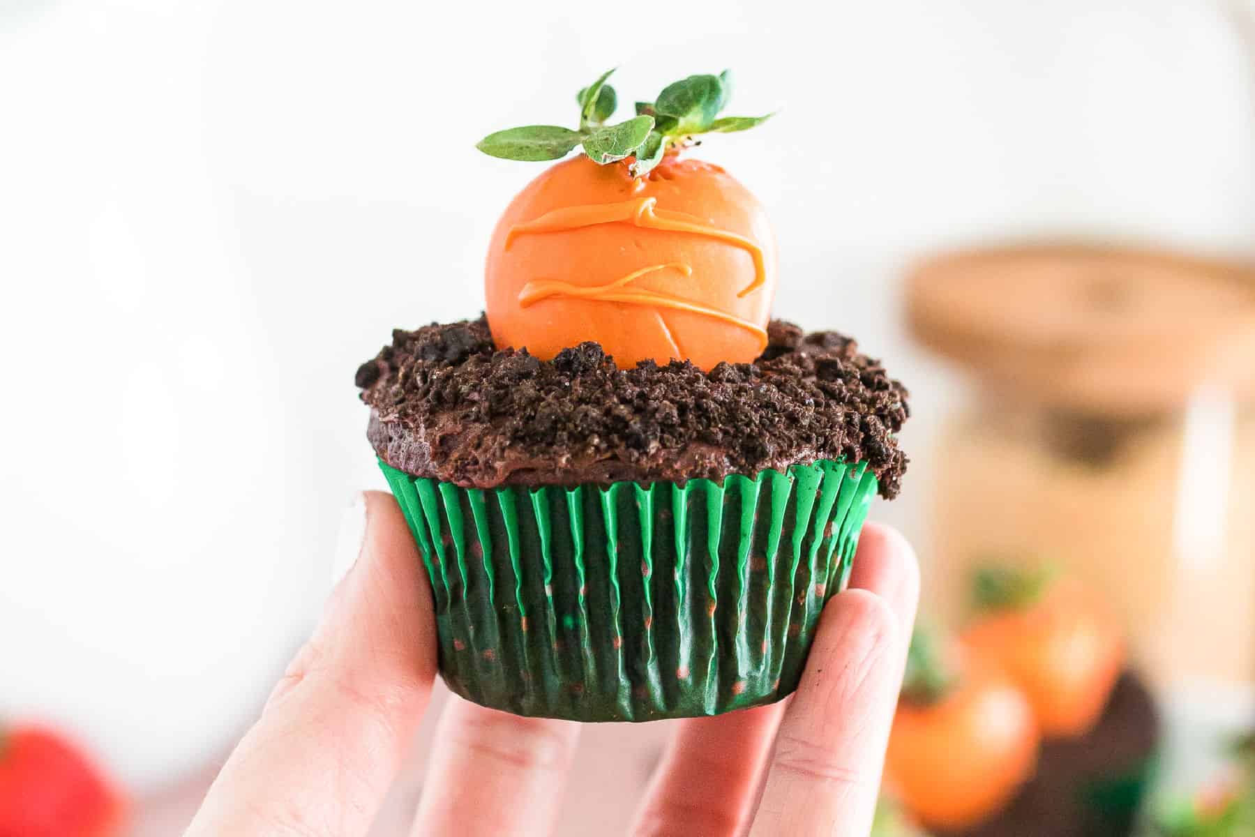 A hand holds a chocolate cupcake with green wrapper, topped with crumbled cookies and an orange chocolate-coated strawberry decorated to look like a carrot—perfect for carrot patch cupcakes.