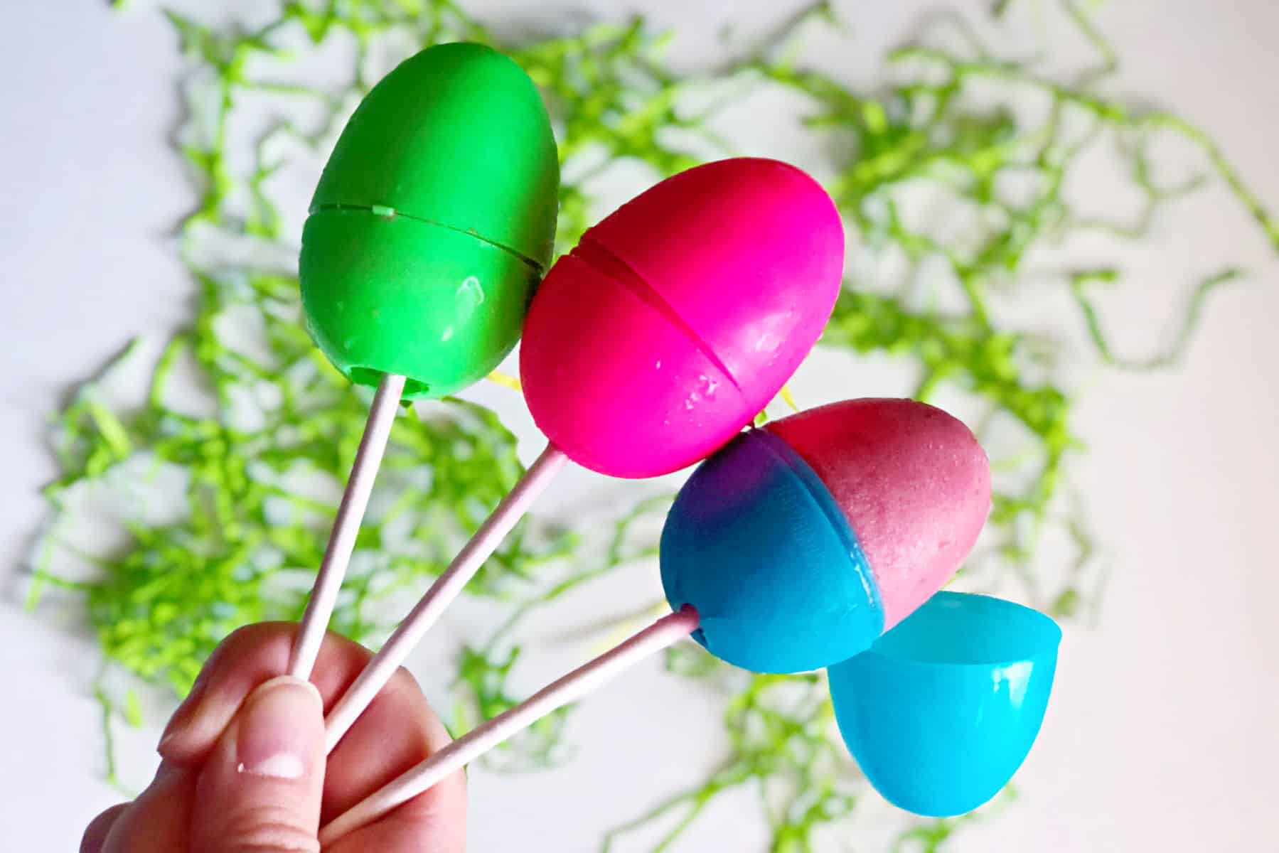 A hand holds three colorful Easter egg popsicles on sticks, with green shredded paper scattered in the background.