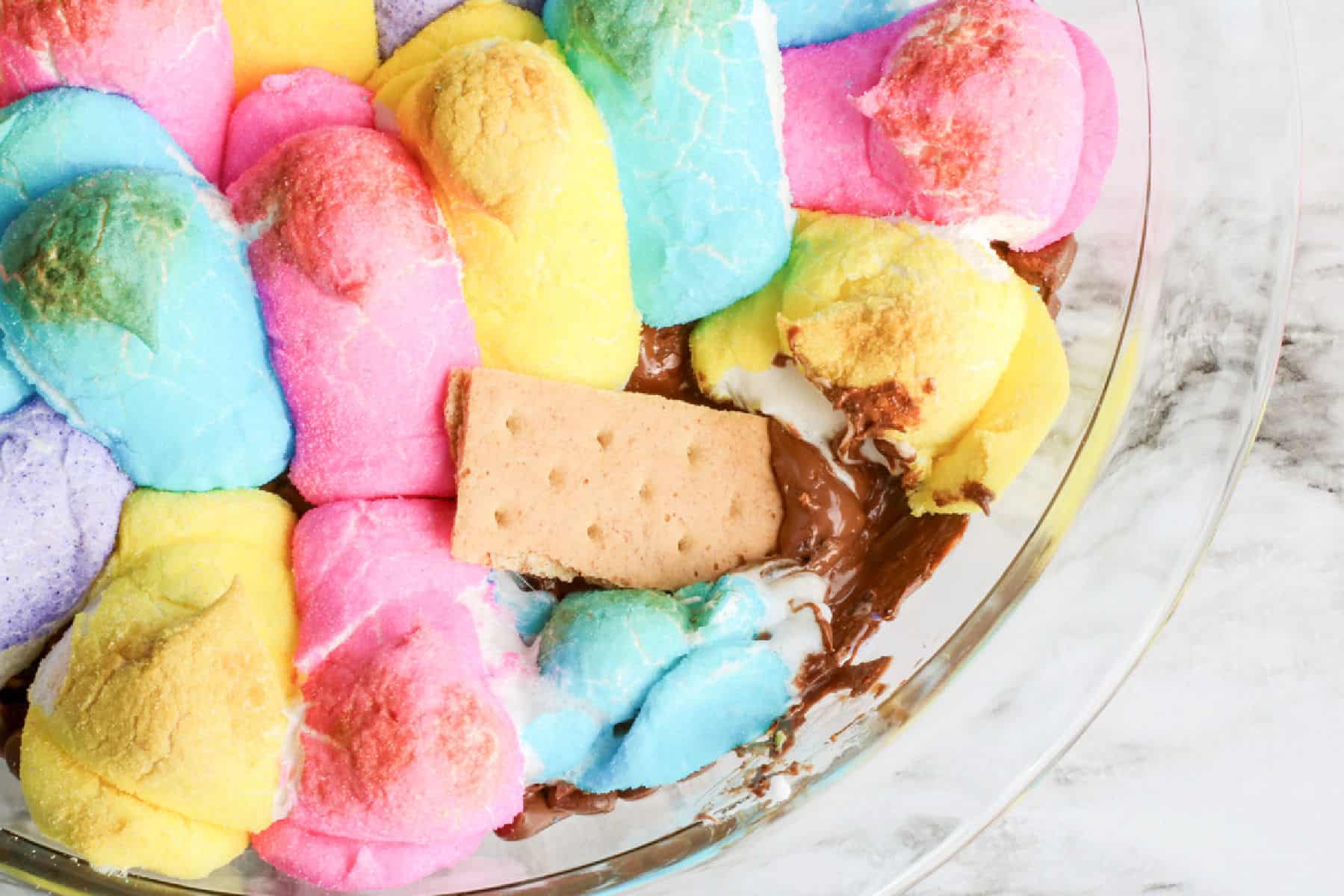A glass dish featuring an Easter Peeps S'mores dip with colorful marshmallow chicks, melted chocolate, and a single graham cracker piece.