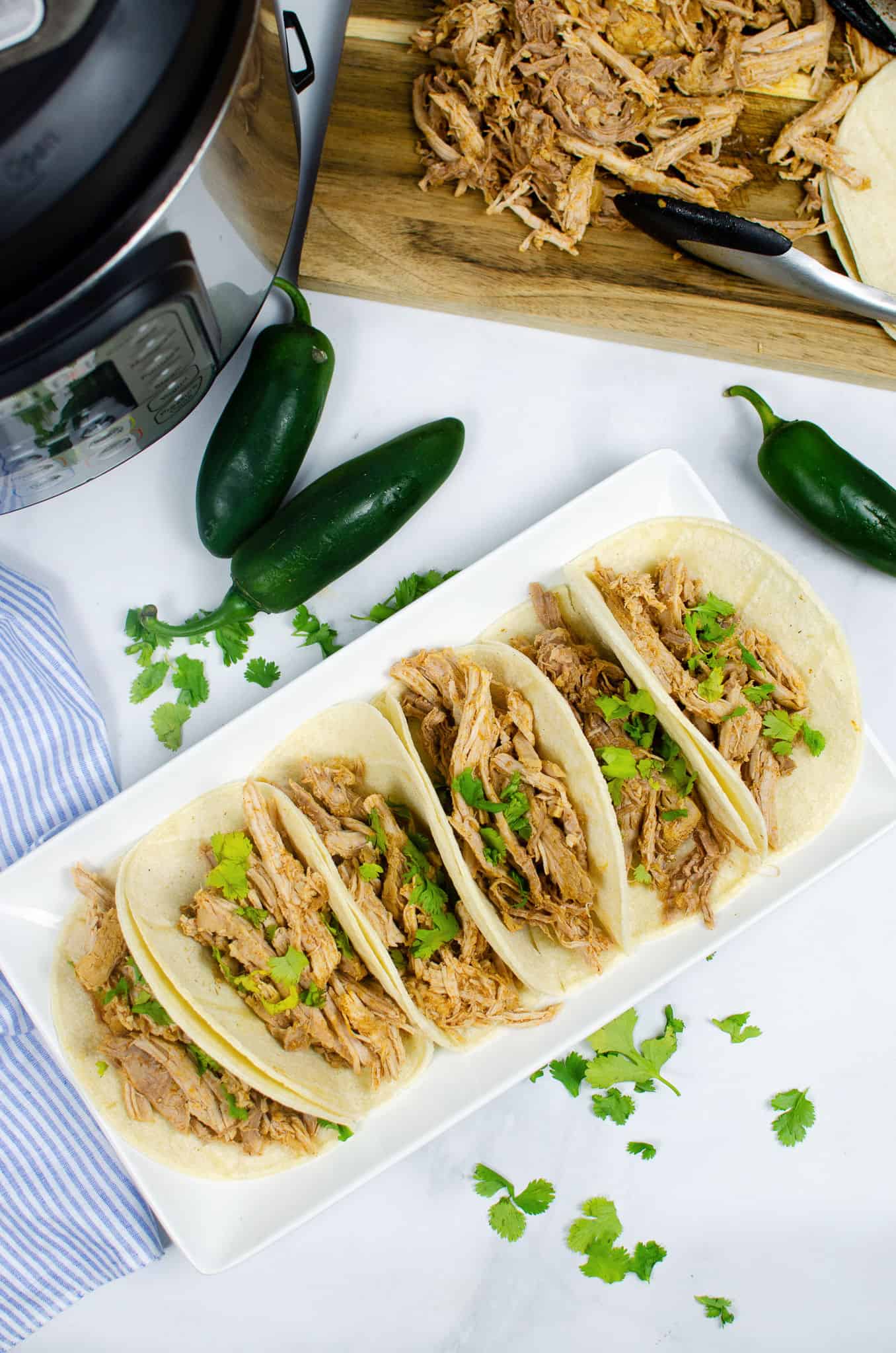 Six instant pot carnitas tacos on a white platter, garnished with cilantro, with jalapeños, cilantro leaves, and extra shredded meat on a cutting board nearby.