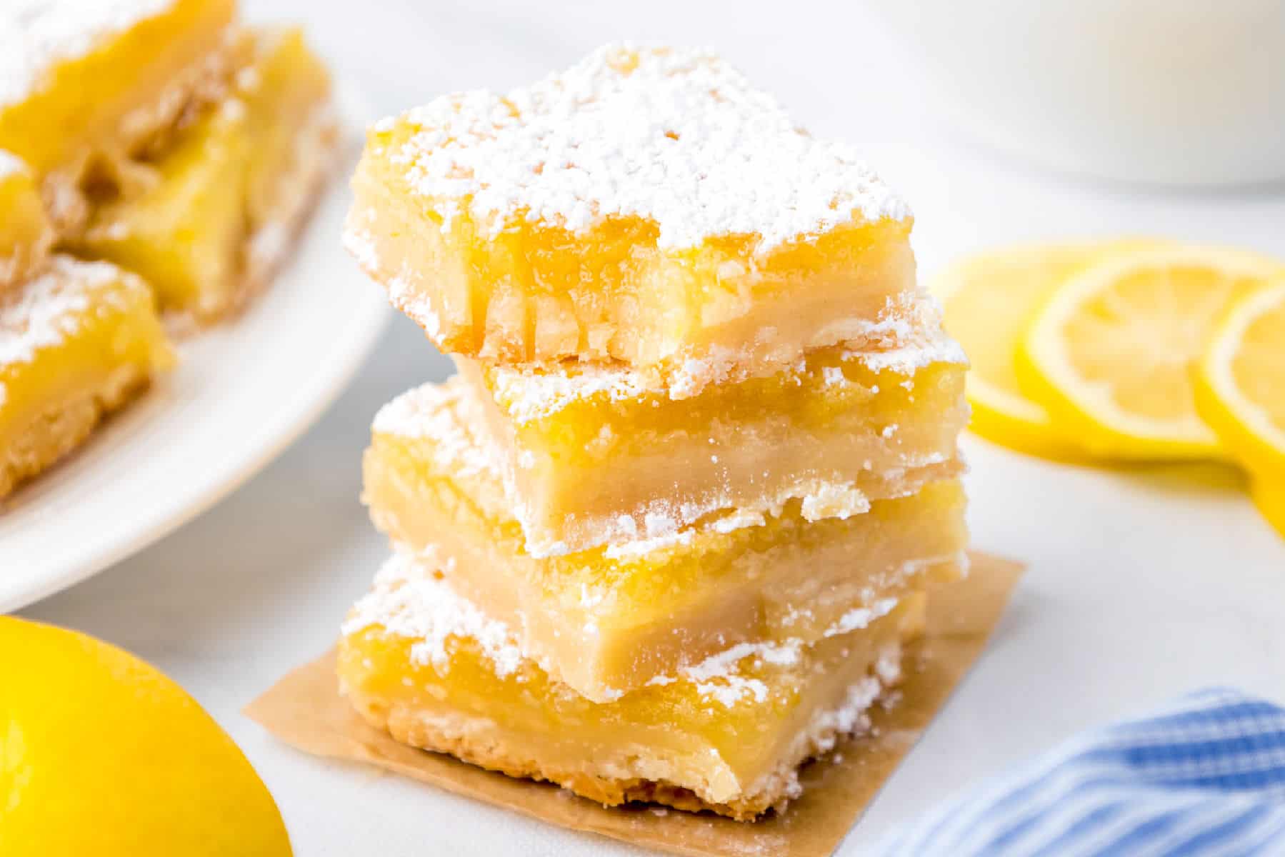 Three lamon bars dusted with powdered sugar are stacked on parchment paper, with lemon slices and more lamon bars visible in the background.