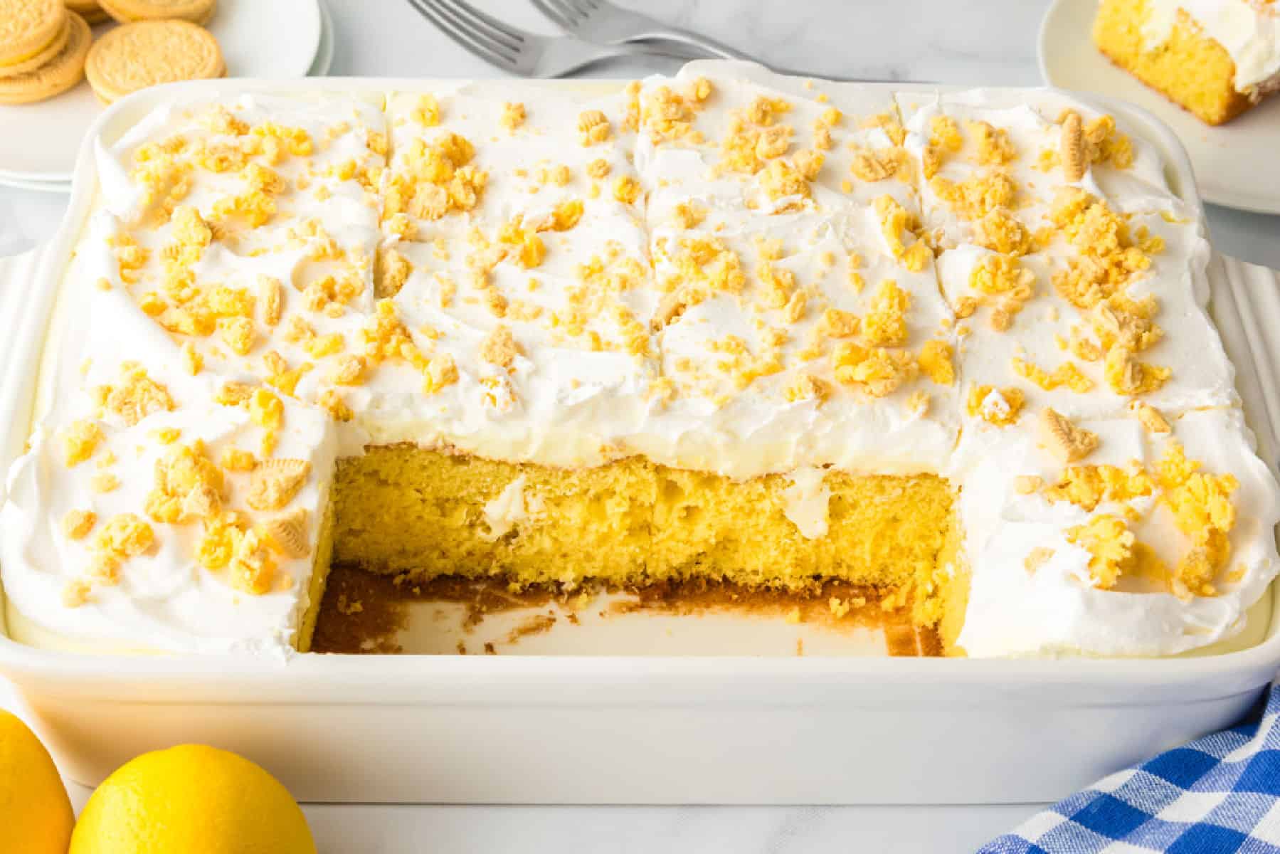 A rectangular lemon pudding poke cake topped with whipped cream and cookie crumbs sits in a white dish with a slice removed; lemons and cookies are nearby.