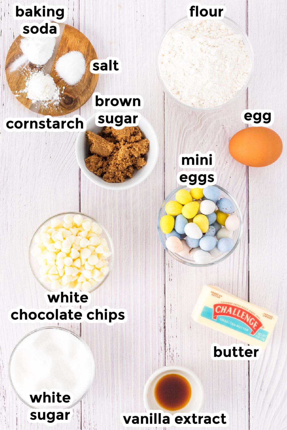 Various ingredients for baking mini egg chocolate chip cookies, including flour, egg, brown sugar, mini eggs, white chocolate chips, butter, vanilla extract, and more are arranged on a white surface.