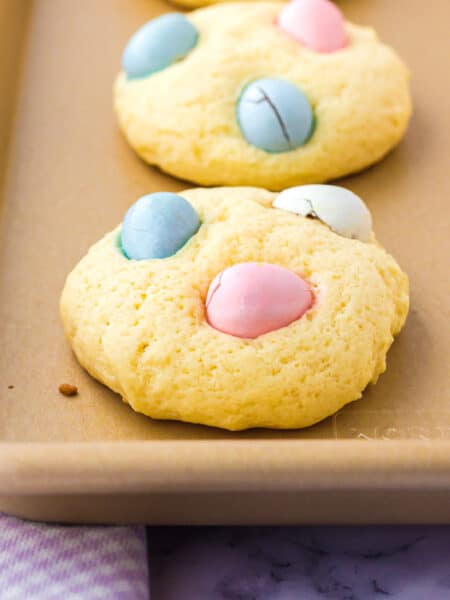 A close-up of mini egg Easter cookies, each topped with pastel-colored candy eggs, on a baking sheet.