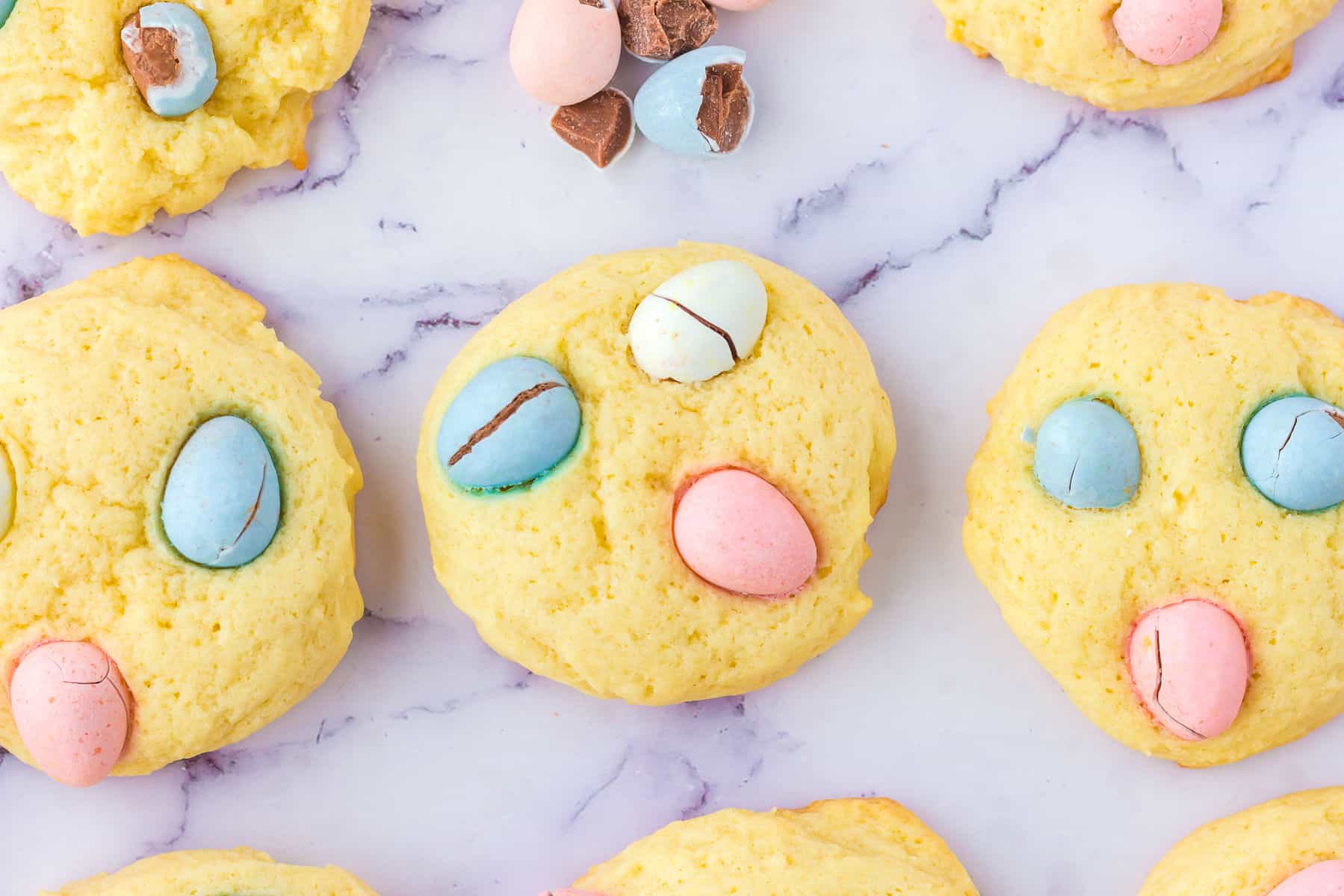 Mini egg sugar cookies topped with pastel-colored candy eggs are arranged on a marble surface, with some broken candies visible.