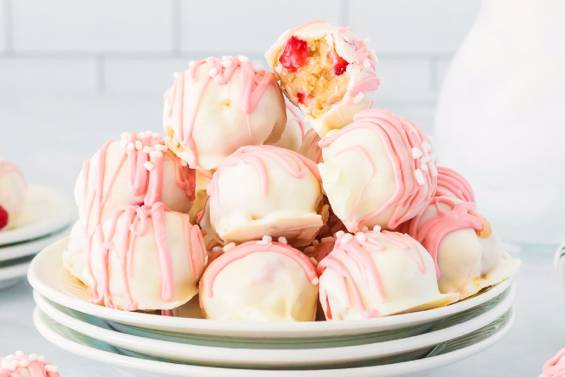 A plate of strawberry shortcake truffles drizzled with pink icing and topped with white sprinkles, with one truffle revealing a luscious cherry filling.
