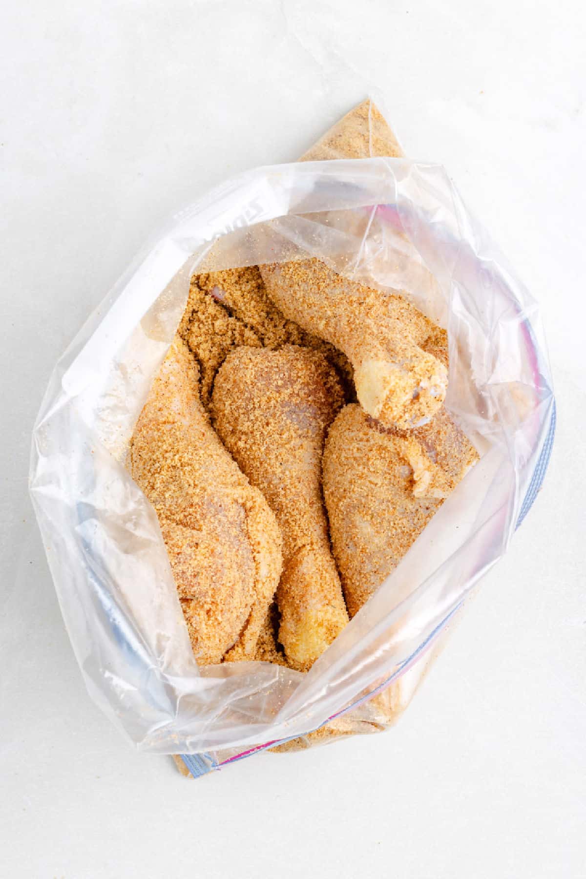 Chicken drumsticks coated in seasoned breading sit inside a partially open plastic zip-top bag, perfect for making shake and bake air fryer chicken drumsticks on a light surface.