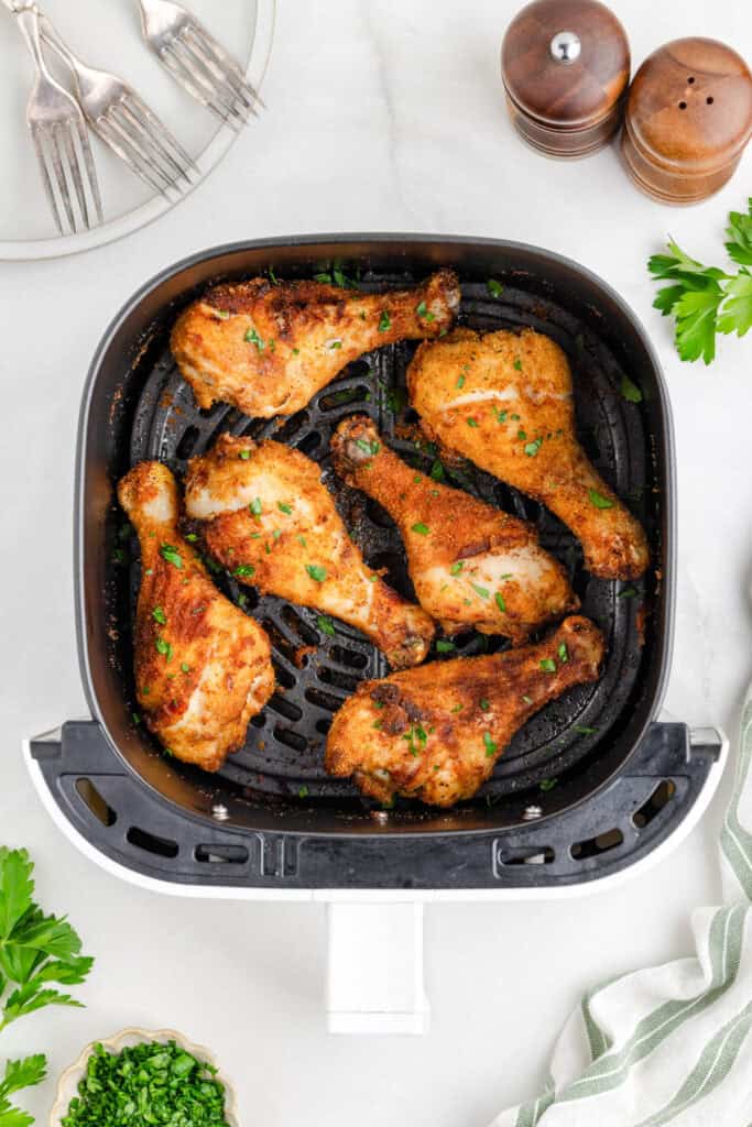 Six seasoned shake and bake air fryer chicken drumsticks cooked in an air fryer basket, garnished with chopped herbs, with plates, forks, salt and pepper shakers, and parsley nearby.