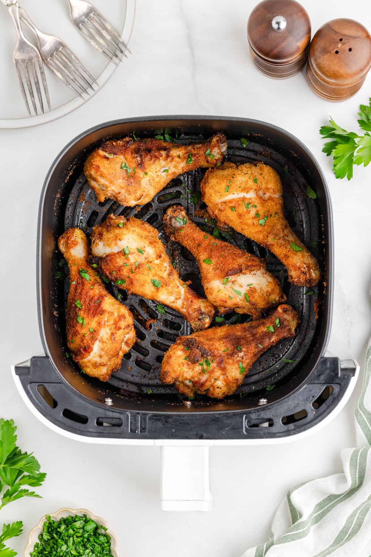 Six seasoned shake and bake air fryer chicken drumsticks cooked in an air fryer basket, garnished with chopped herbs, with plates, forks, salt and pepper shakers, and parsley nearby.