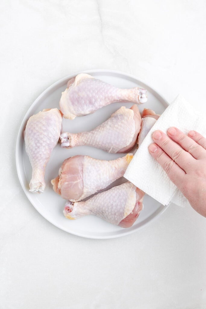 A hand uses a paper towel to pat dry raw chicken drumsticks arranged on a white plate, prepping them for delicious shake and bake air fryer chicken drumsticks.