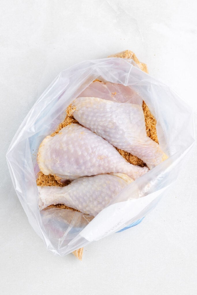Four raw chicken drumsticks coated with seasoning are placed inside a clear plastic bag on a white surface, ready to become shake and bake air fryer chicken drumsticks.