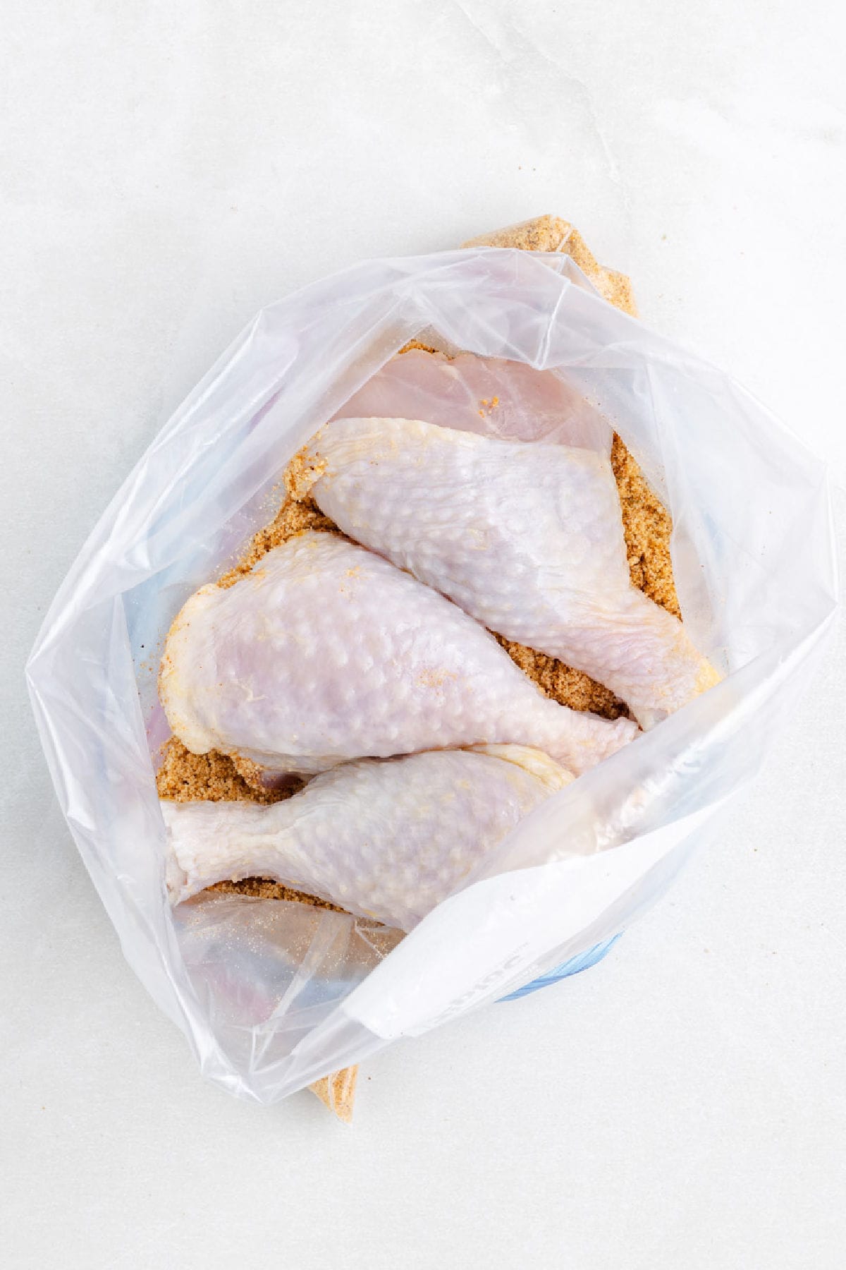Four raw chicken drumsticks coated with seasoning are placed inside a clear plastic bag on a white surface, ready to become shake and bake air fryer chicken drumsticks.