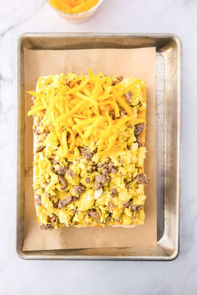 A sheet pan with the bottom of slider rolls topped with scrambled eggs, cooked ground sausage, half covered in shredded cheddar cheese for breakfast sliders.