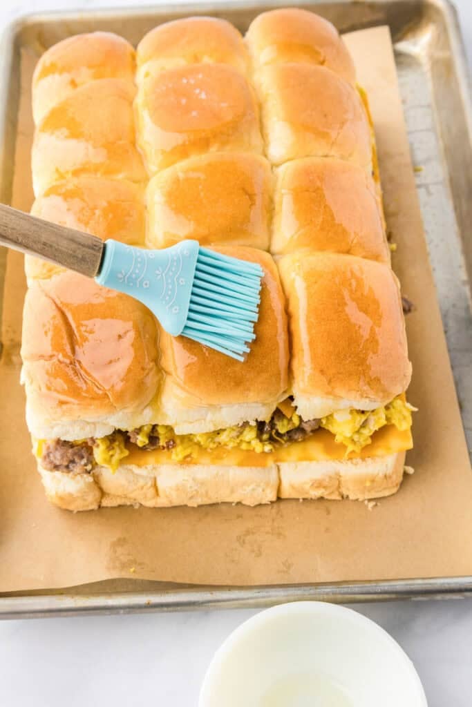 A silicone brush is spreading melted butter on top of a batch of breakfast sliders, filled with eggs, sausage, and cheese, all arranged neatly on a baking sheet lined with parchment paper.