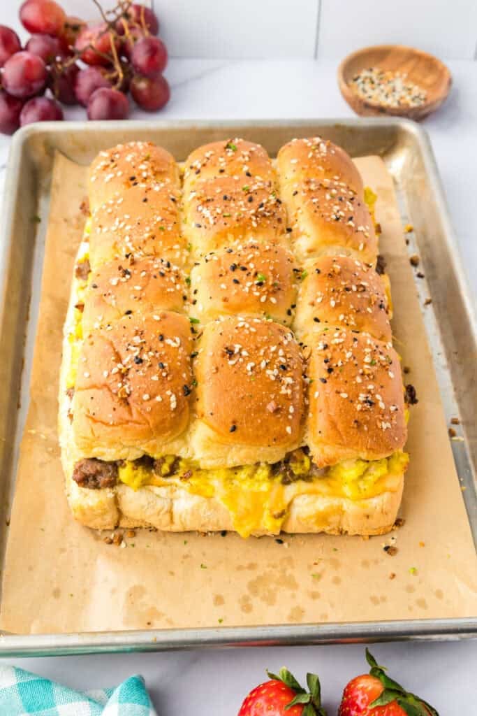A tray of baked breakfast sliders made with dinner rolls, scrambled eggs, and sausage, topped with seasoning, sits on parchment paper.