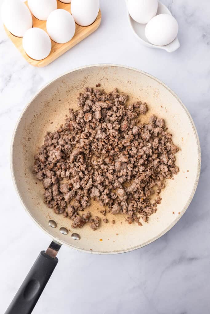A frying pan with cooked ground sausage sits on a marble countertop, ready to be layered into breakfast sliders, with a tray and bowl of whole eggs nearby.