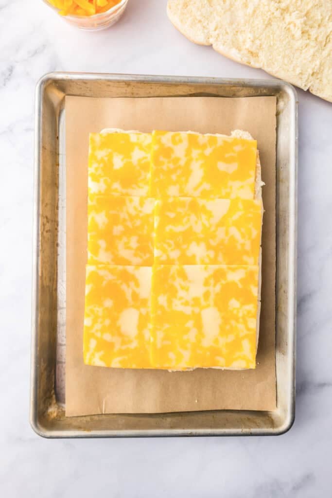 A baking sheet lined with parchment paper holds sandwich bread topped with six slices of marbled yellow and white cheese, perfect for making delicious breakfast sliders.