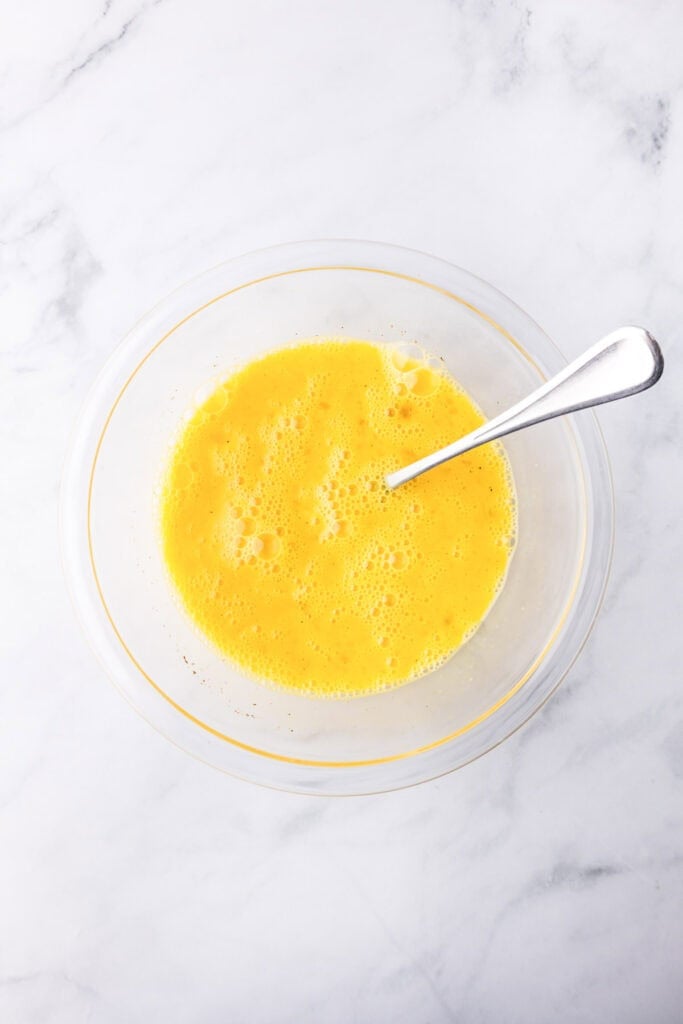 A clear glass bowl filled with beaten eggs and a metal fork on a white marble surface, perfect for preparing breakfast sliders.