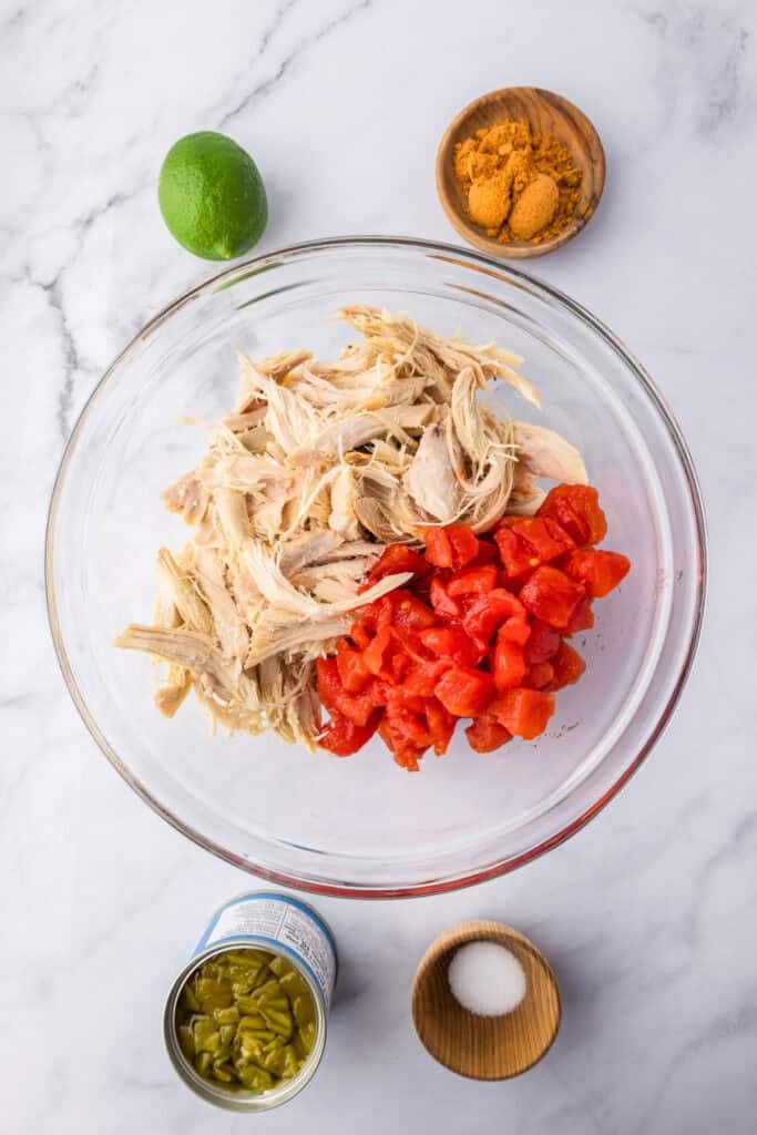 A glass bowl with shredded chicken and diced tomatoes to make the filling for crispy baked chicken tacos, with other ingredients on the counter nearby.