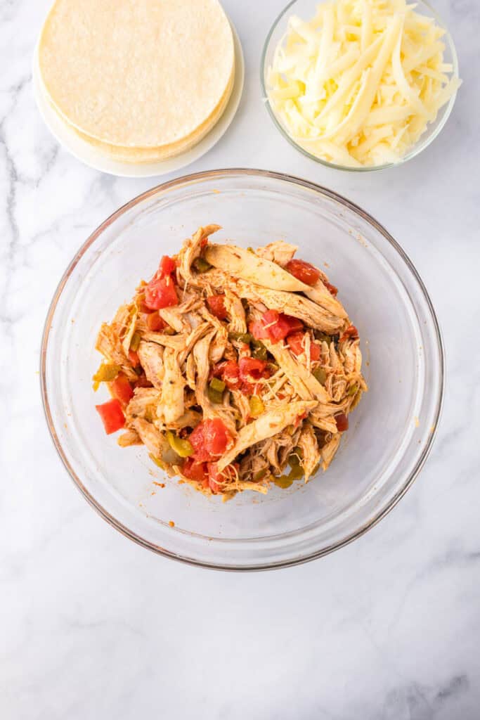 A glass bowl of shredded chicken with tomatoes and peppers for crispy baked chicken tacos, with corn tortillas and cheese nearby on the counter.